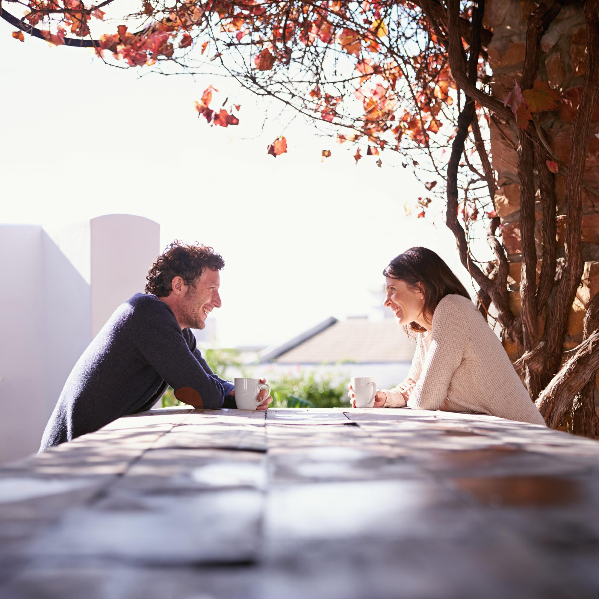 A couple sitting under autumn trees at a wooden table, smiling and talking, reflecting emotional connection and growth at a couples retreat.