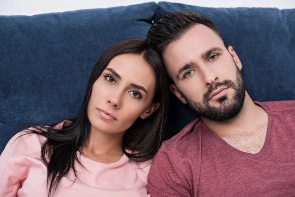 Couple relaxing on couch, man and woman leaning close together, looking serious.