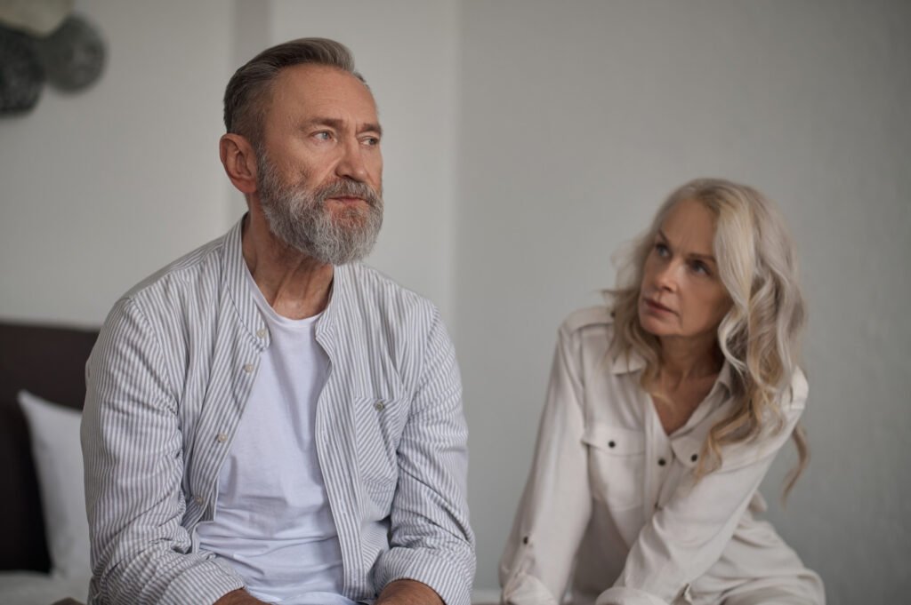 An older couple sitting together after an argument, with the husband looking away as his wife tries to engage, symbolizes the withdrawal and detachment of a covert narcissist father.