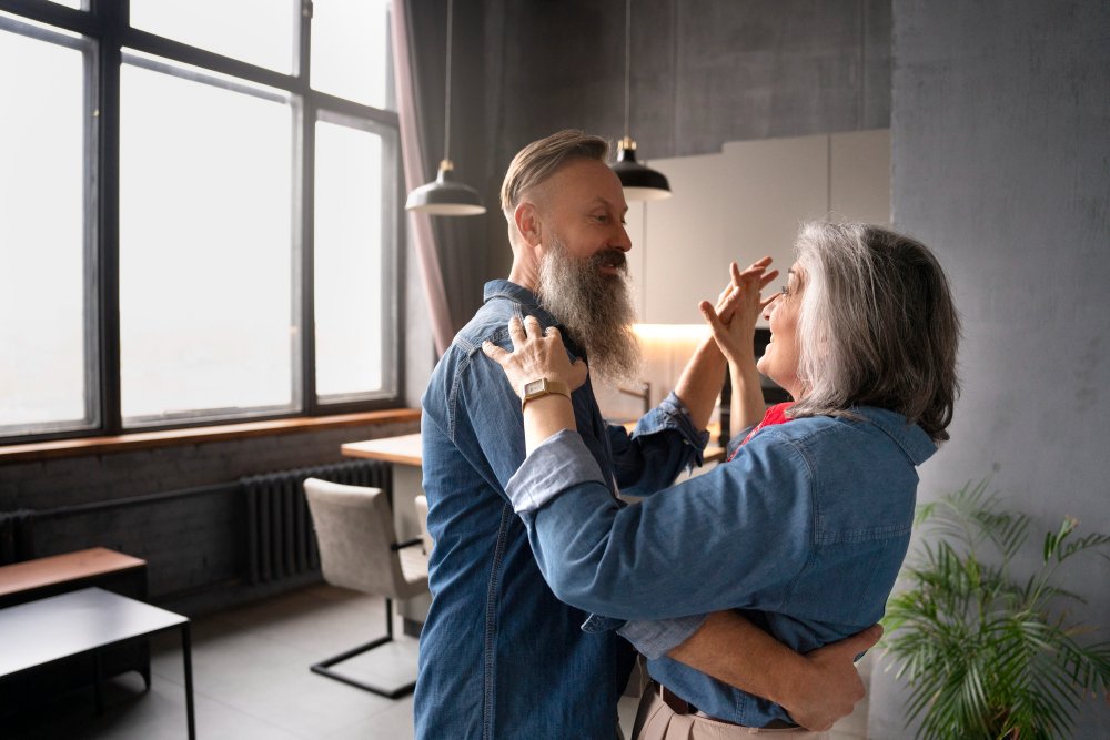  An older man and woman smile warmly as they dance together in a bright, modern room, holding each other with relaxed, joyful expressions.