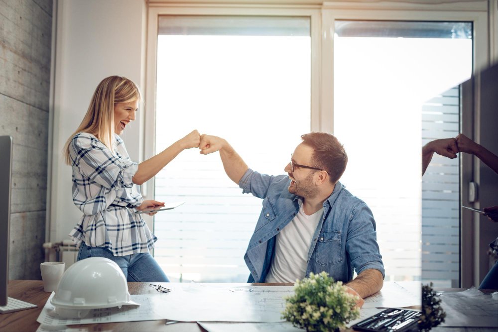 A smiling woman and man enthusiastically bump fists across a desk during a lighthearted moment in a bright office setting.
