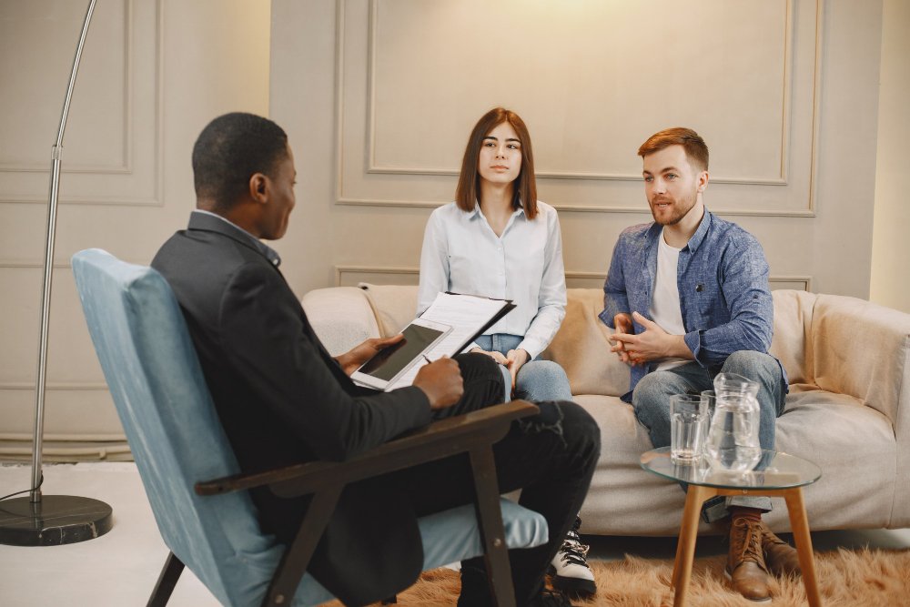 Couple seated together in a therapy session with a therapist taking notes, reflecting, and guided discussion typical of intensive couples therapy work
