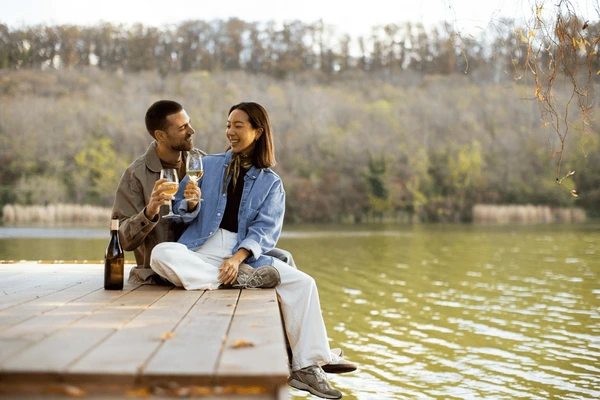 Romantic couple enjoying white wine on a wooden dock by a peaceful lake surrounded by autumn trees.
