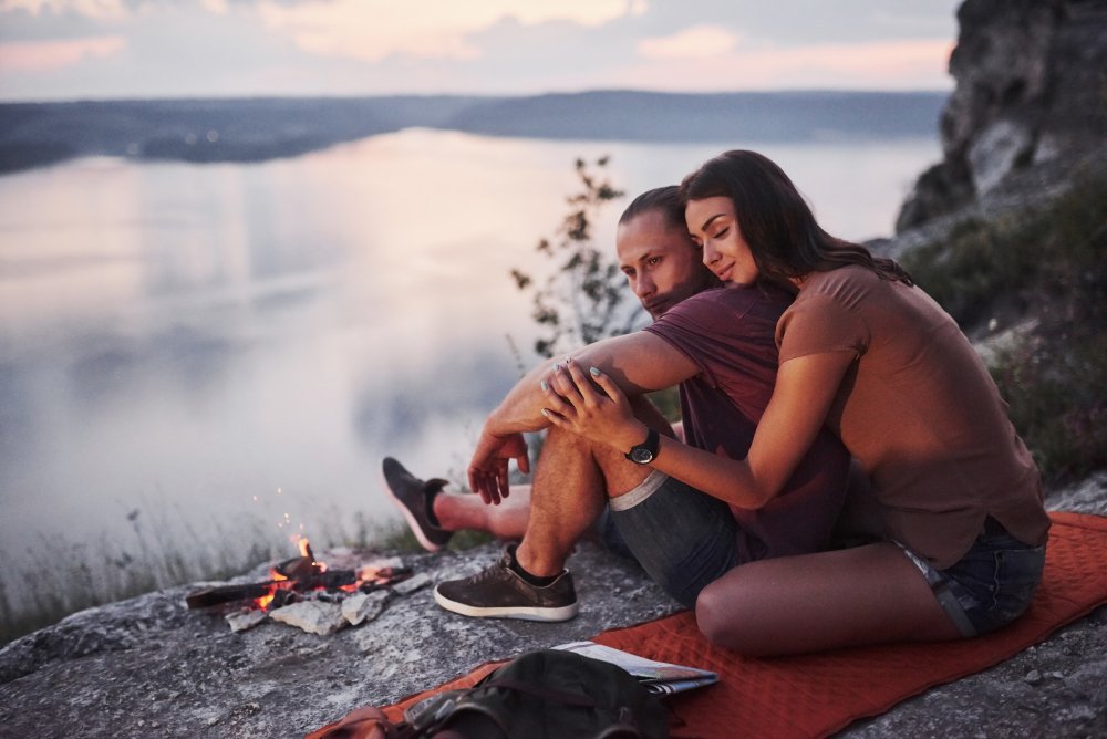 Hugging couple with backpacks relaxing by a campfire on a mountain, overlooking a lake on their retreat.