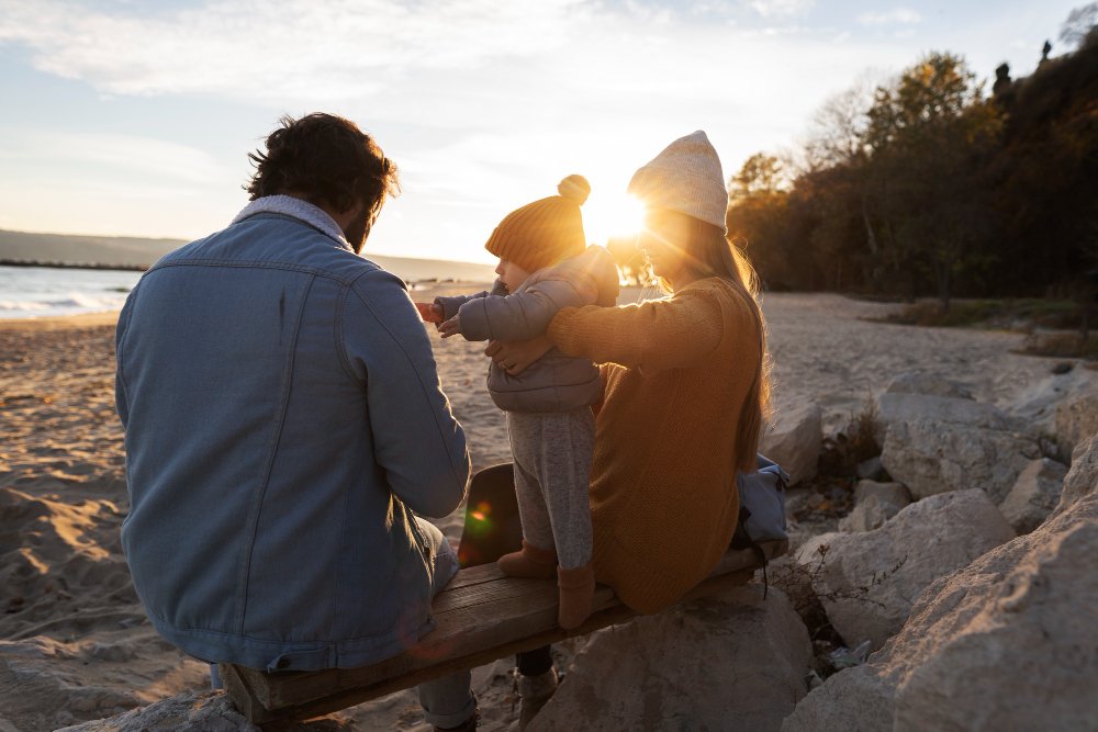 A father and a mother sitting on the rocks at the beach with their child at sunset.