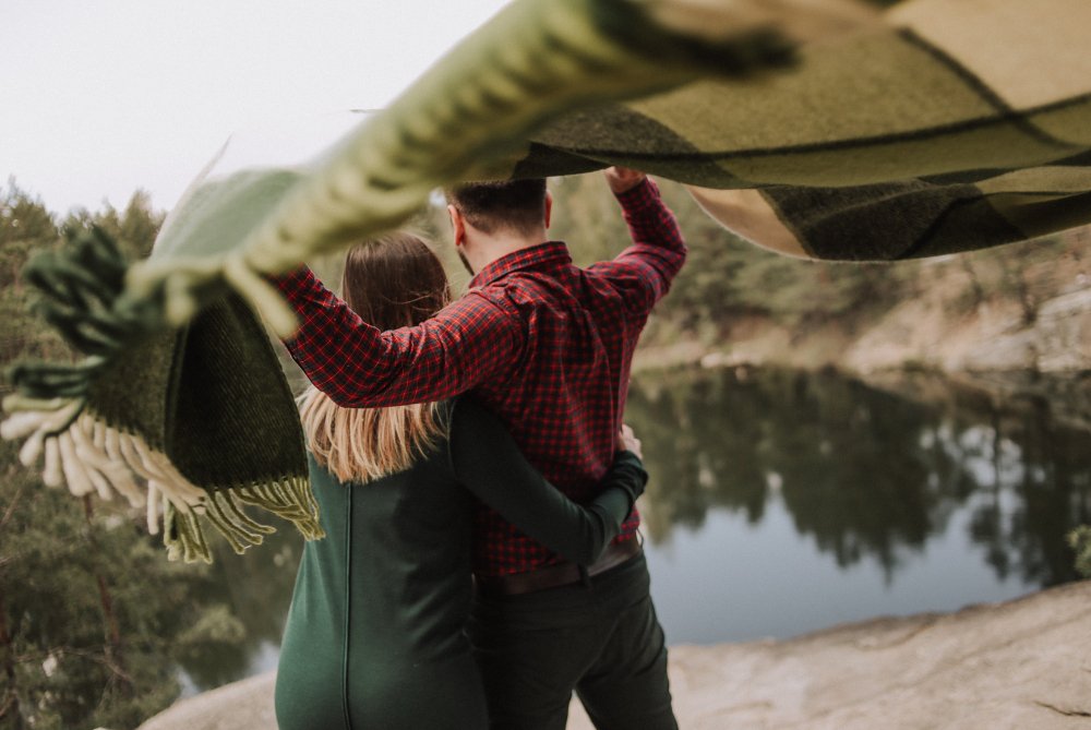 Couple wrapped in a blanket by a lake shore with a steep cliff, sharing a quiet retreat moment. 