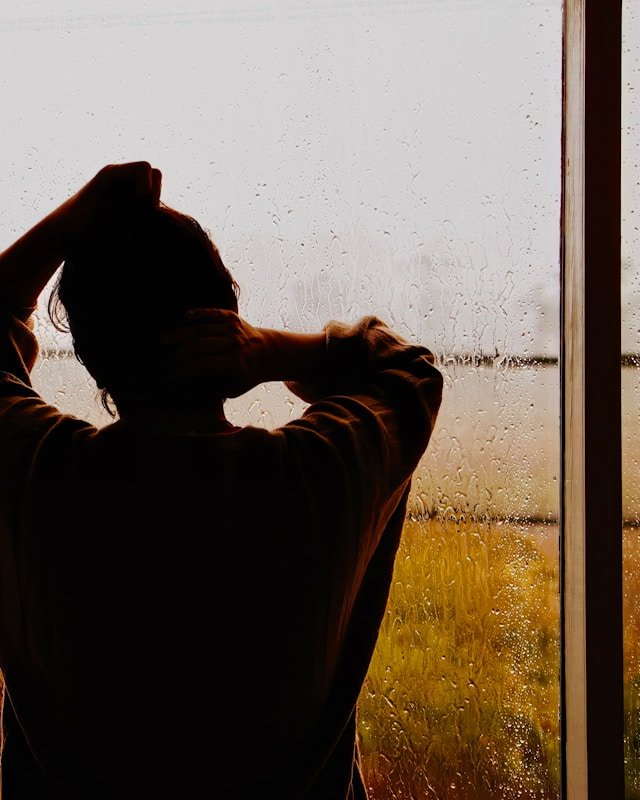 A man stands alone at a rain-streaked window reflecting on the intrusive thoughts and isolation that follow infidelity trauma 
